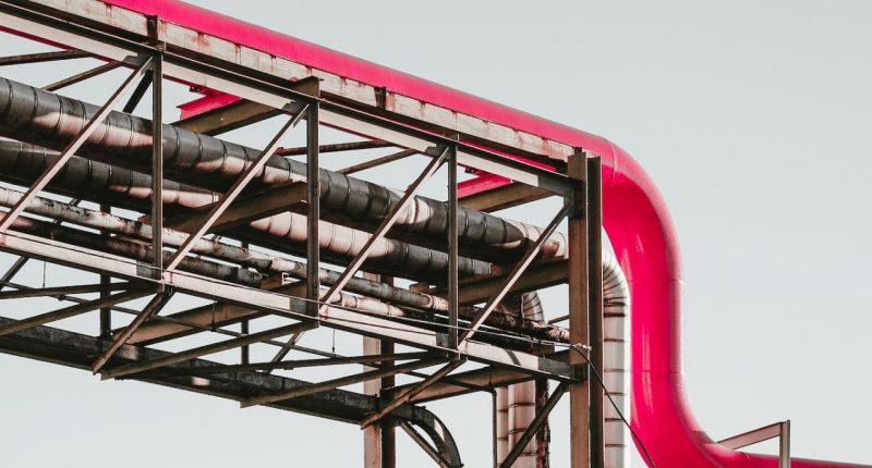 A pipeline, owner unknown, runs across the screen. A grey scaffolding structure holds up a newer added pipe in pink.