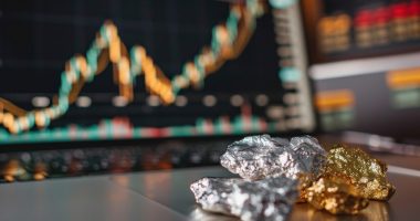 Raw chunks of silver and gold in focus with a blurred stock market graph on a computer monitor in the backdrop