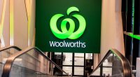 A big green Woolworths logo and sign hanging above shopping market escalators.