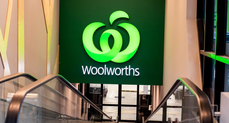 A big green Woolworths logo and sign hanging above shopping market escalators.