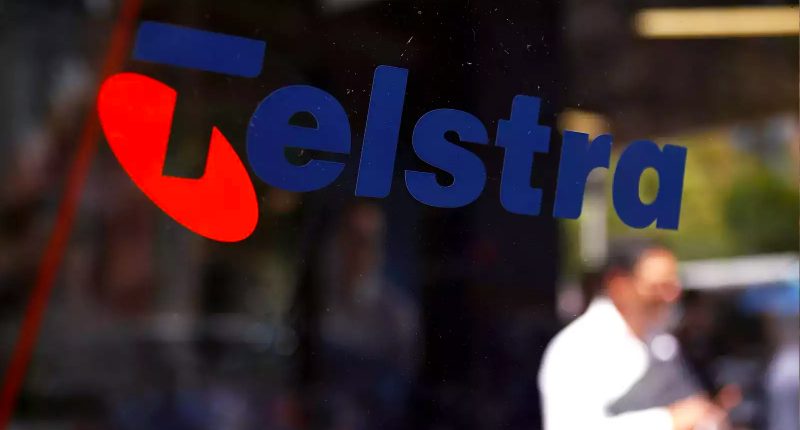 A Telstra logo on the side of a window. You can see a man on his phone in the background.