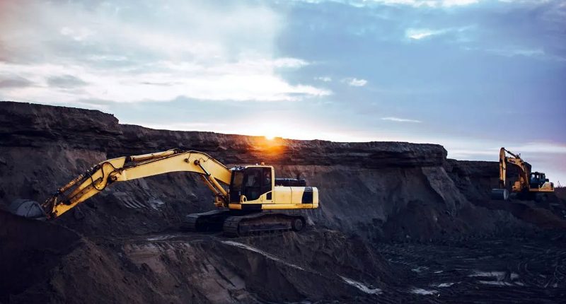A large yellow digger working on deep dark dirt under a cloudy sky.