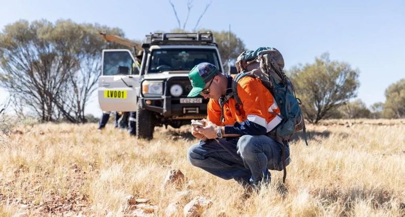 A worker in high-vis safety gear examines a machine on the ground in a field. A truck is parked behind him with an open door.