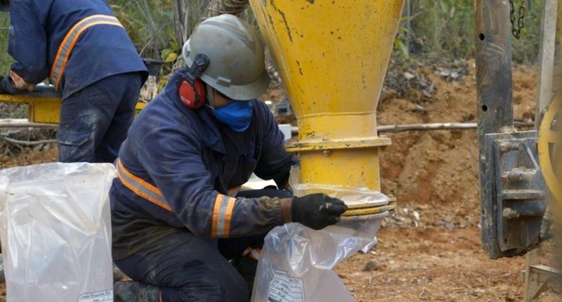 A worker fits a plastic bag over the end of a yellow funnel's nozzle, which is facing the chipper dirt.