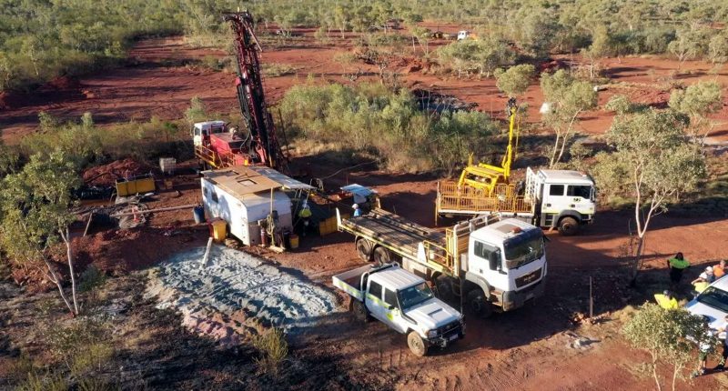 Trucks and a rig at the Cloncurry Gold Project. There's bushes and dirt around the worksite.