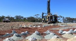 Bags of soil samples littered across a heaped field. A big gold rig sits in the background.
