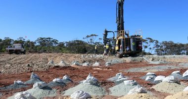 Bags of soil samples littered across a heaped field. A big gold rig sits in the background.