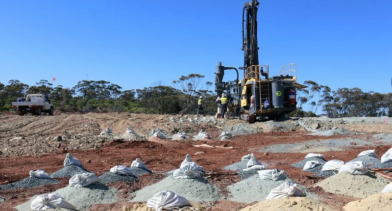 Bags of soil samples littered across a heaped field. A big gold rig sits in the background.