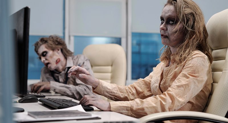 Two zombie works sit at an office desk.